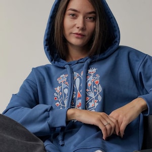 May include: A blue hooded sweatshirt with a colorful embroidered design on the front. The design features geometric shapes and floral motifs in red, orange, and white.