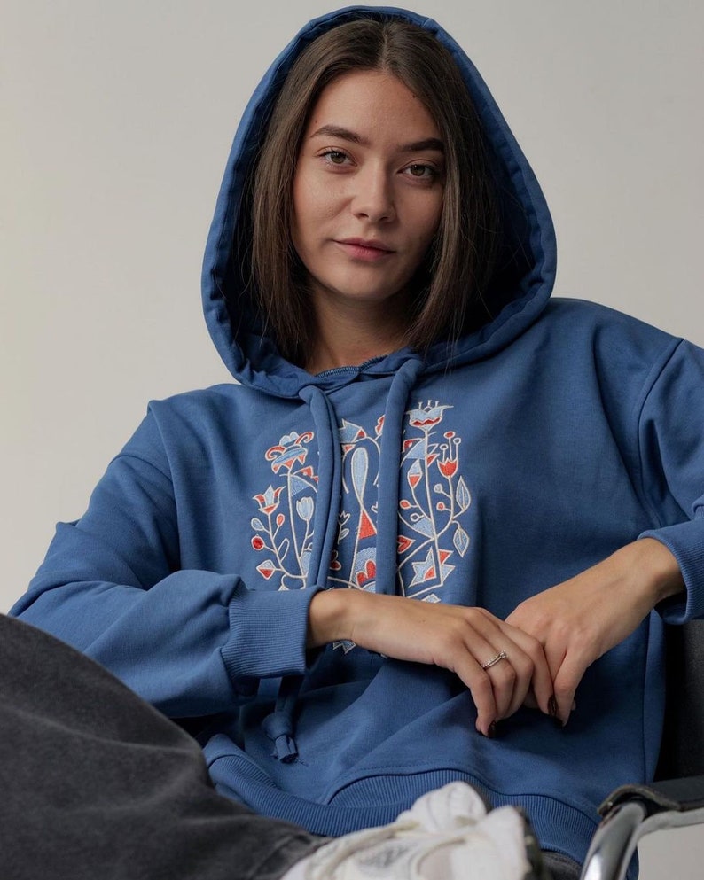 May include: A blue hooded sweatshirt with a colorful embroidered design on the front. The design features geometric shapes and floral motifs in red, orange, and white.