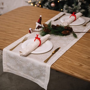 May include: A white table runner with a snowflake pattern, featuring two place settings with white plates and napkins. Each napkin is held by a red reindeer napkin ring.