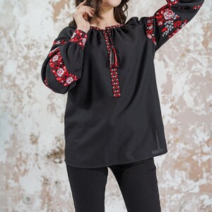 May include: A black embroidered blouse with red and white floral patterns on the sleeves. The blouse has a round neckline and a tassel detail at the front.