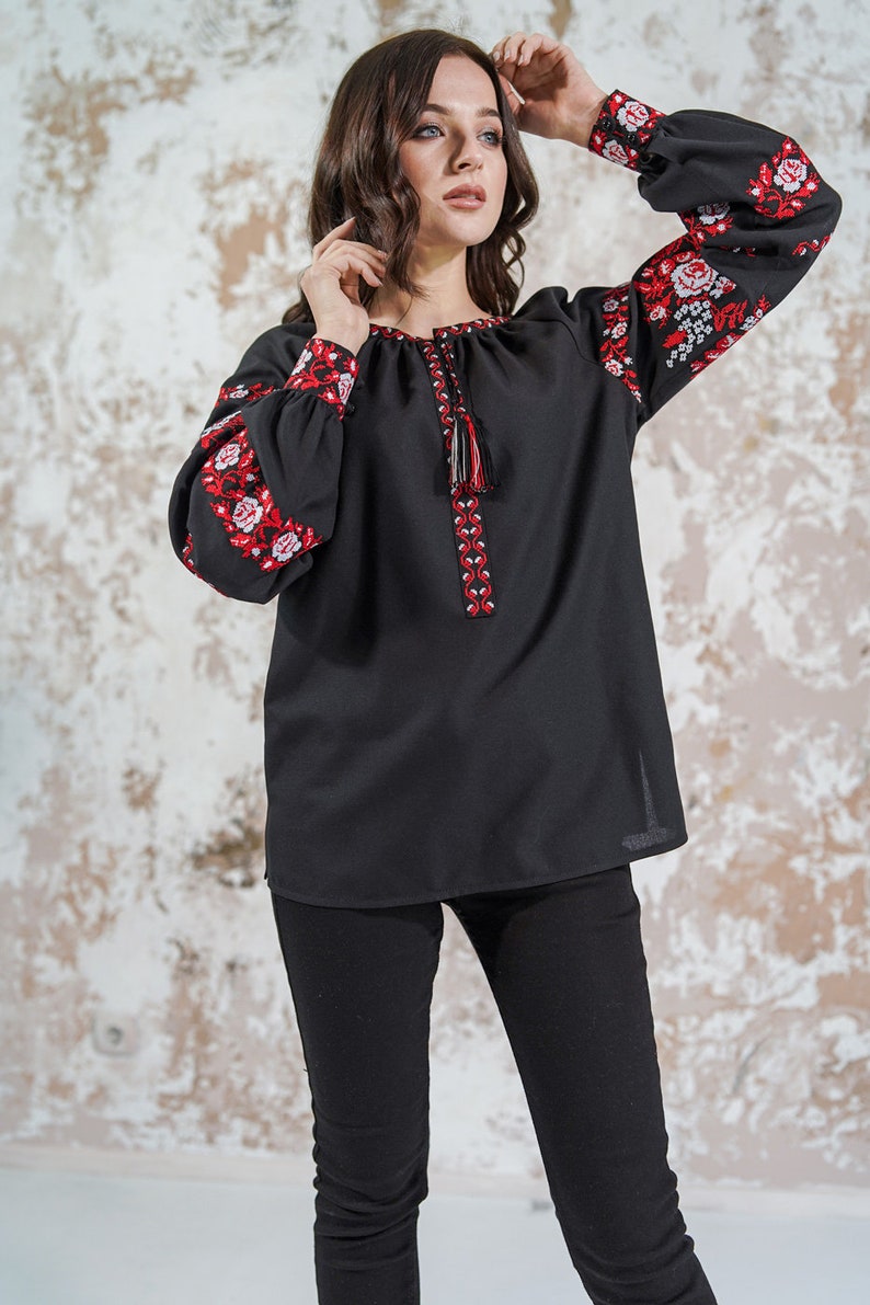 May include: A black embroidered blouse with red and white floral patterns on the sleeves. The blouse has a round neckline and a tassel detail at the front.