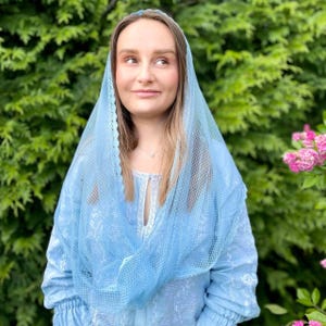 May include: A light blue veil and matching long-sleeved top. The veil has a delicate, scalloped edge and a sheer, patterned design. The top features a keyhole neckline with white lace trim and gathered sleeves. The background is a lush green with pink flowers.