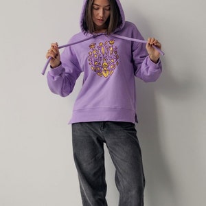 May include: A lavender hoodie with a yellow and purple embroidered design on the front. The design features a stylized floral pattern.