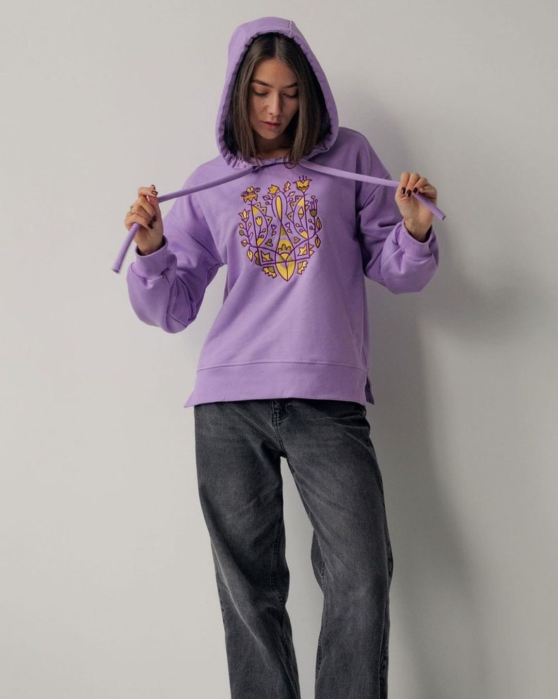 May include: A lavender hoodie with a yellow and purple embroidered design on the front. The design features a stylized floral pattern.