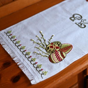 以下が含まれることがあります： 白いリネンのイースターテーブルランナー。刺繍されたイースターエッグと柳の枝が描かれています。卵は赤と緑の模様で装飾されています。端には刺繍のボーダーとフリンジが付いています。