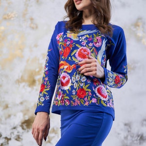 May include: A two-piece outfit in cobalt blue, comprising a long-sleeved top and a skirt. The top is adorned with colourful floral embroidery, including red, pink, and yellow flowers, and a bird motif. The skirt is a plain, matching blue.