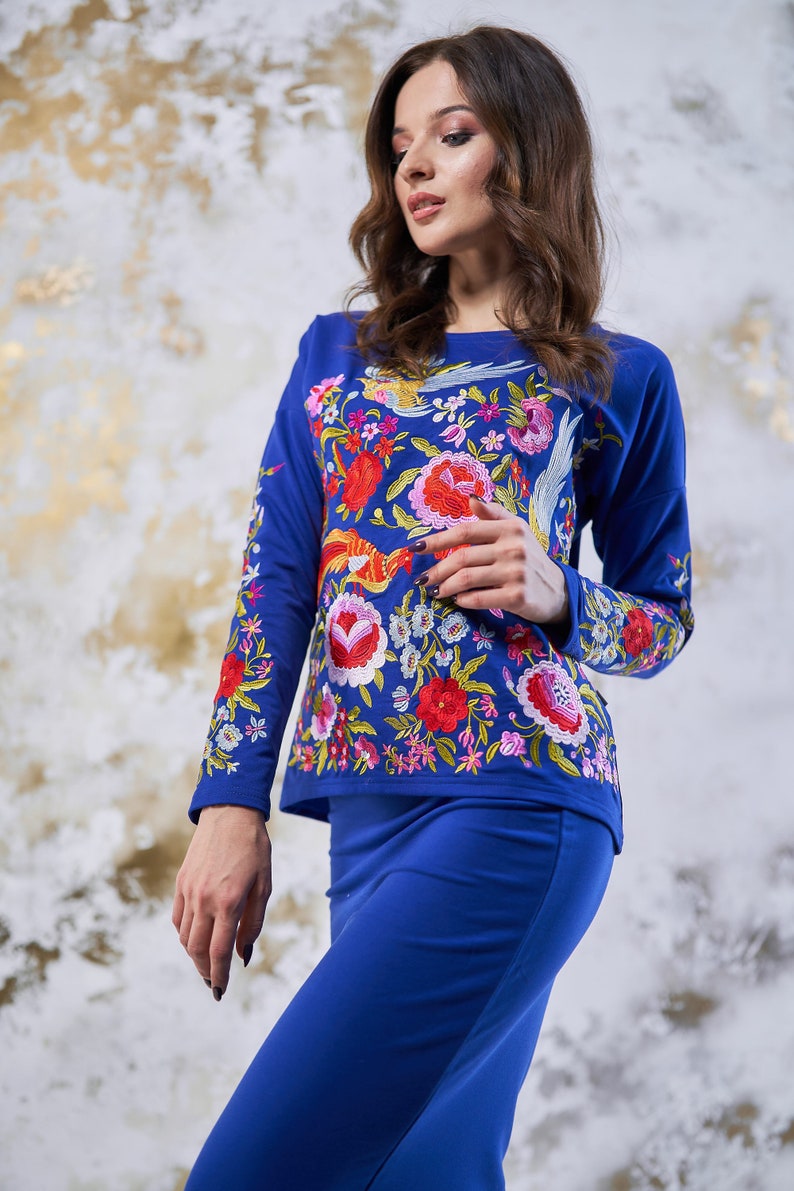 May include: A two-piece outfit in cobalt blue, comprising a long-sleeved top and a skirt. The top is adorned with colourful floral embroidery, including red, pink, and yellow flowers, and a bird motif. The skirt is a plain, matching blue.