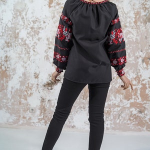 May include: A black embroidered blouse with red and white floral designs on the sleeves. The blouse has a gathered neckline and long sleeves. The model is wearing black pants and high heels.