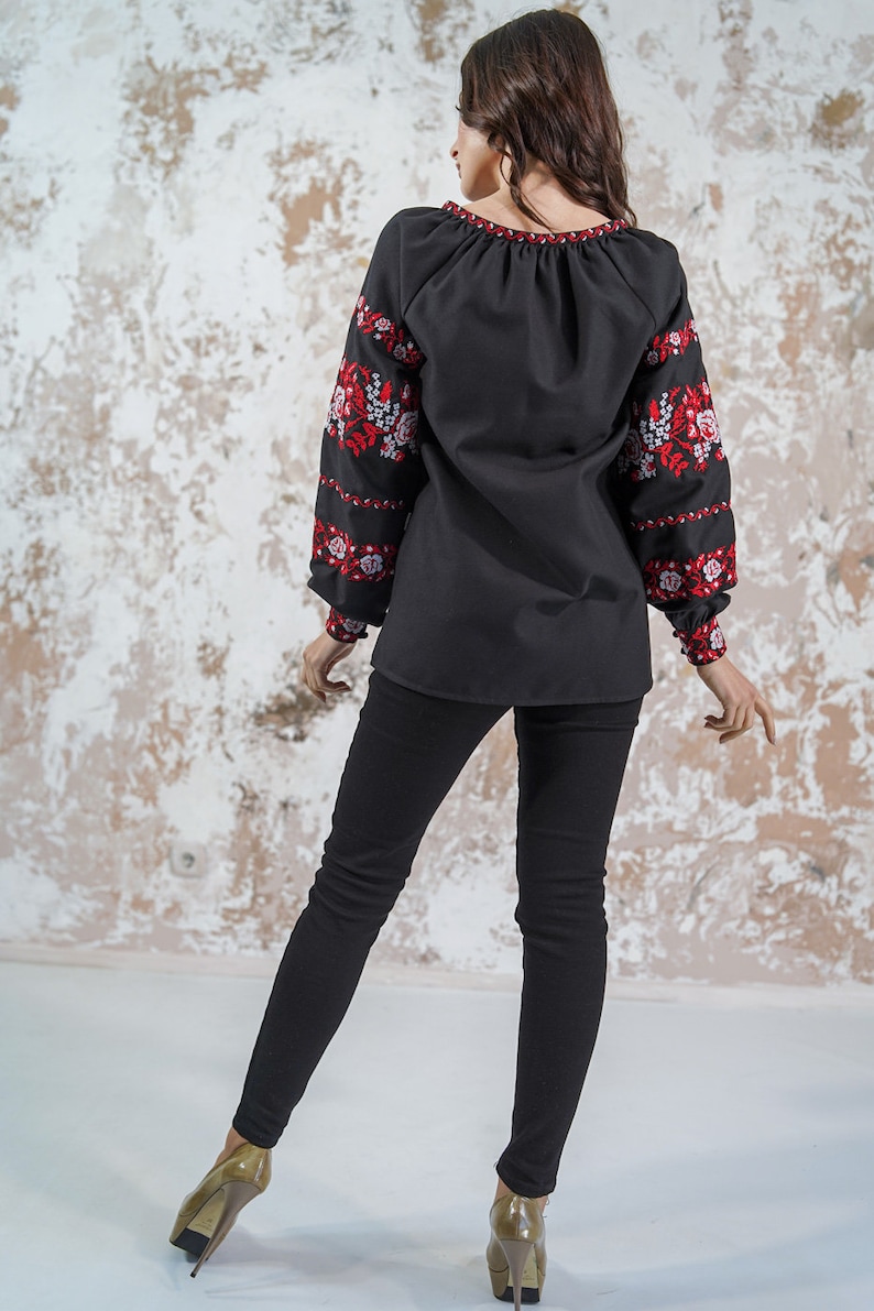 May include: A black embroidered blouse with red and white floral designs on the sleeves. The blouse has a gathered neckline and long sleeves. The model is wearing black pants and high heels.