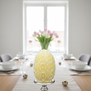 K&ouml;nnte beinhalten: Ein gro&szlig;es, gelbes Osterei mit wei&szlig;er Verzierung, pr&auml;sentiert auf einem silbernen Sockel. Das Ei ist der Mittelpunkt eines Esstisches, mit einer Vase mit rosa und wei&szlig;en Tulpen im Hintergrund.