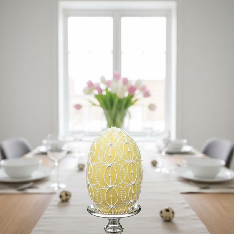 K&ouml;nnte beinhalten: Ein gro&szlig;es, gelbes Osterei mit wei&szlig;er Verzierung, pr&auml;sentiert auf einem silbernen Sockel. Das Ei ist der Mittelpunkt eines Esstisches, mit einer Vase mit rosa und wei&szlig;en Tulpen im Hintergrund.