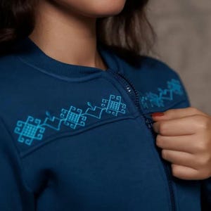 Puede incluir: Una sudadera azul oscuro con cremallera y un dise&ntilde;o bordado azul claro en el hombro. El dise&ntilde;o presenta formas geom&eacute;tricas.