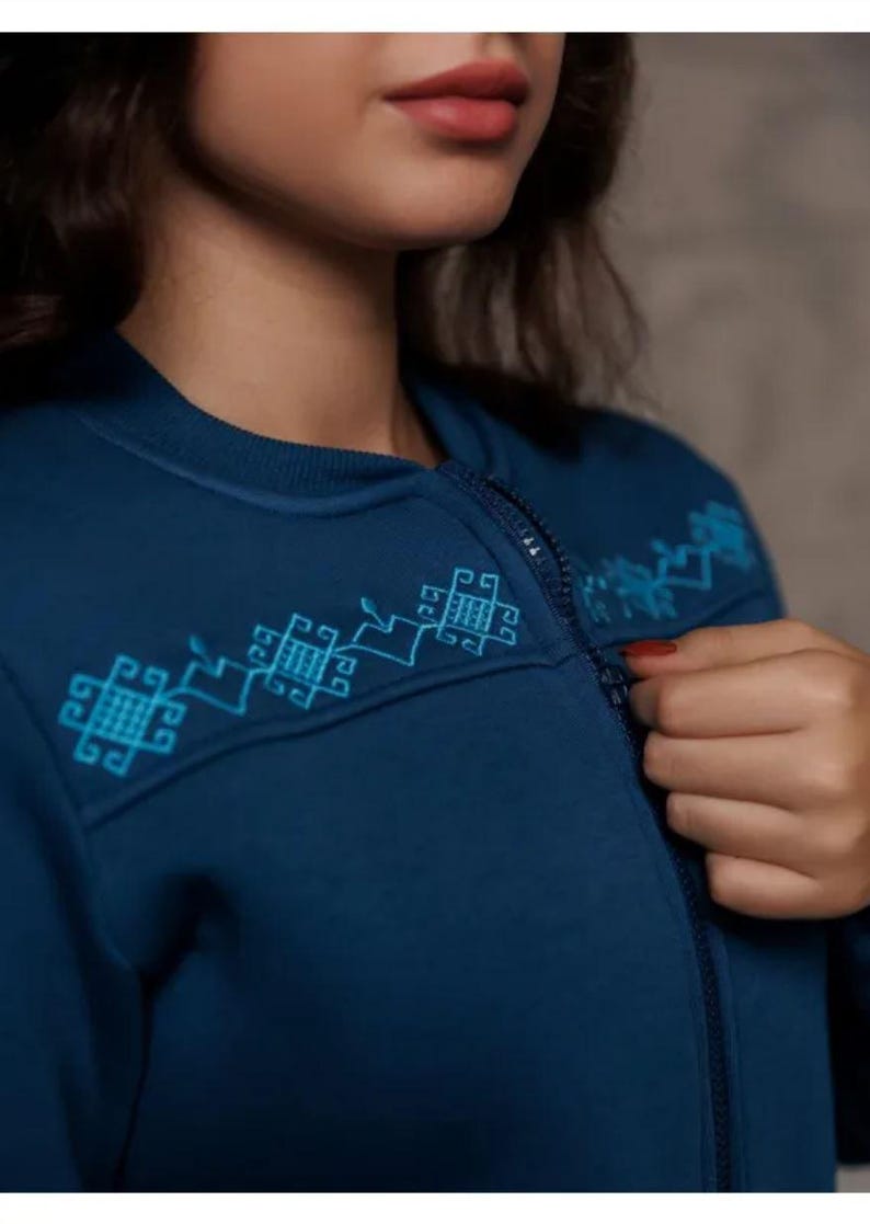 Puede incluir: Una sudadera azul oscuro con cremallera y un dise&ntilde;o bordado azul claro en el hombro. El dise&ntilde;o presenta formas geom&eacute;tricas.
