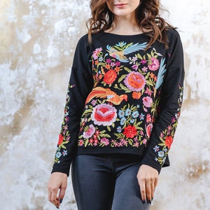 May include: Black long-sleeved top with colourful floral and bird embroidery. The embroidery includes red, orange, pink, blue, and green. The top is worn with dark grey jeans.