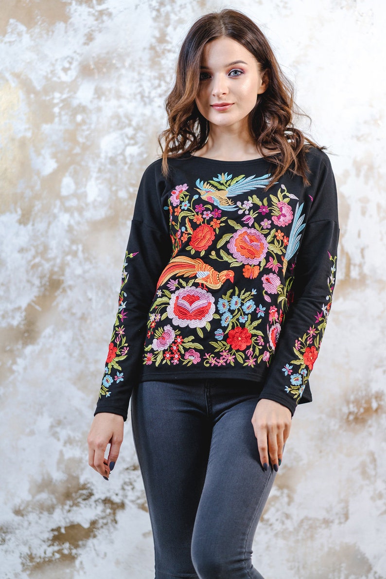 May include: Black long-sleeved top with colourful floral and bird embroidery. The embroidery includes red, orange, pink, blue, and green. The top is worn with dark grey jeans.