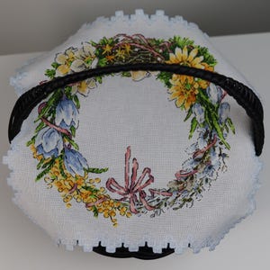 Könnte beinhalten: Ein schwarzer Weidenkorb mit einem weißen Stofffutter, das mit einem floralen Kranz verziert ist. Der Kranz besteht aus blauen und gelben Blumen, grünen Blättern und rosa Bändern.