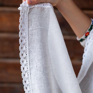 Puede incluir: Primer plano de una tela blanca texturizada con un delicado ribete de encaje. La tela se sostiene, mostrando los intrincados detalles del encaje y el tejido del material. El fondo es una superficie de madera.