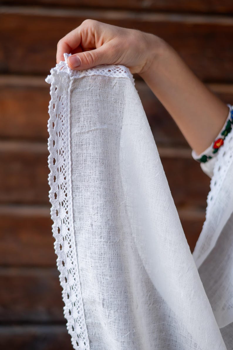 Puede incluir: Primer plano de una tela blanca texturizada con un delicado ribete de encaje. La tela se sostiene, mostrando los intrincados detalles del encaje y el tejido del material. El fondo es una superficie de madera.