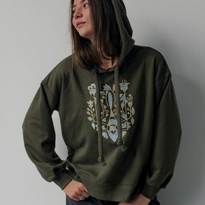 May include: A dark green hooded sweatshirt with a floral design in shades of gold, blue, and brown. The design features stylized flowers and leaves.