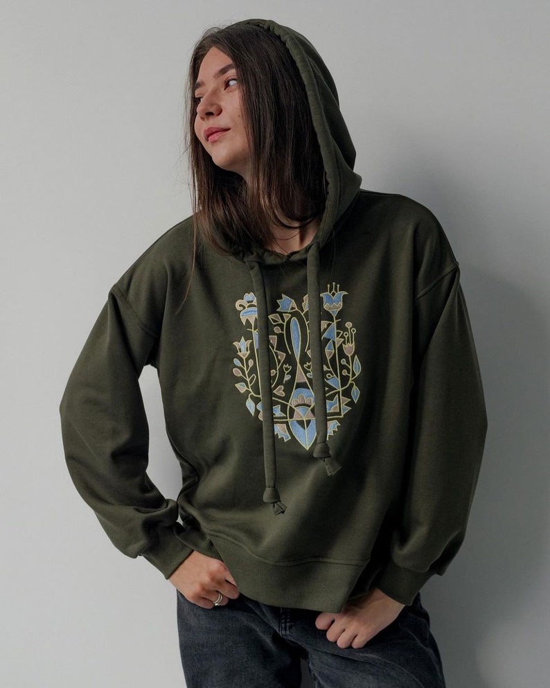 May include: A dark green hooded sweatshirt with a floral design in shades of gold, blue, and brown. The design features stylized flowers and leaves.