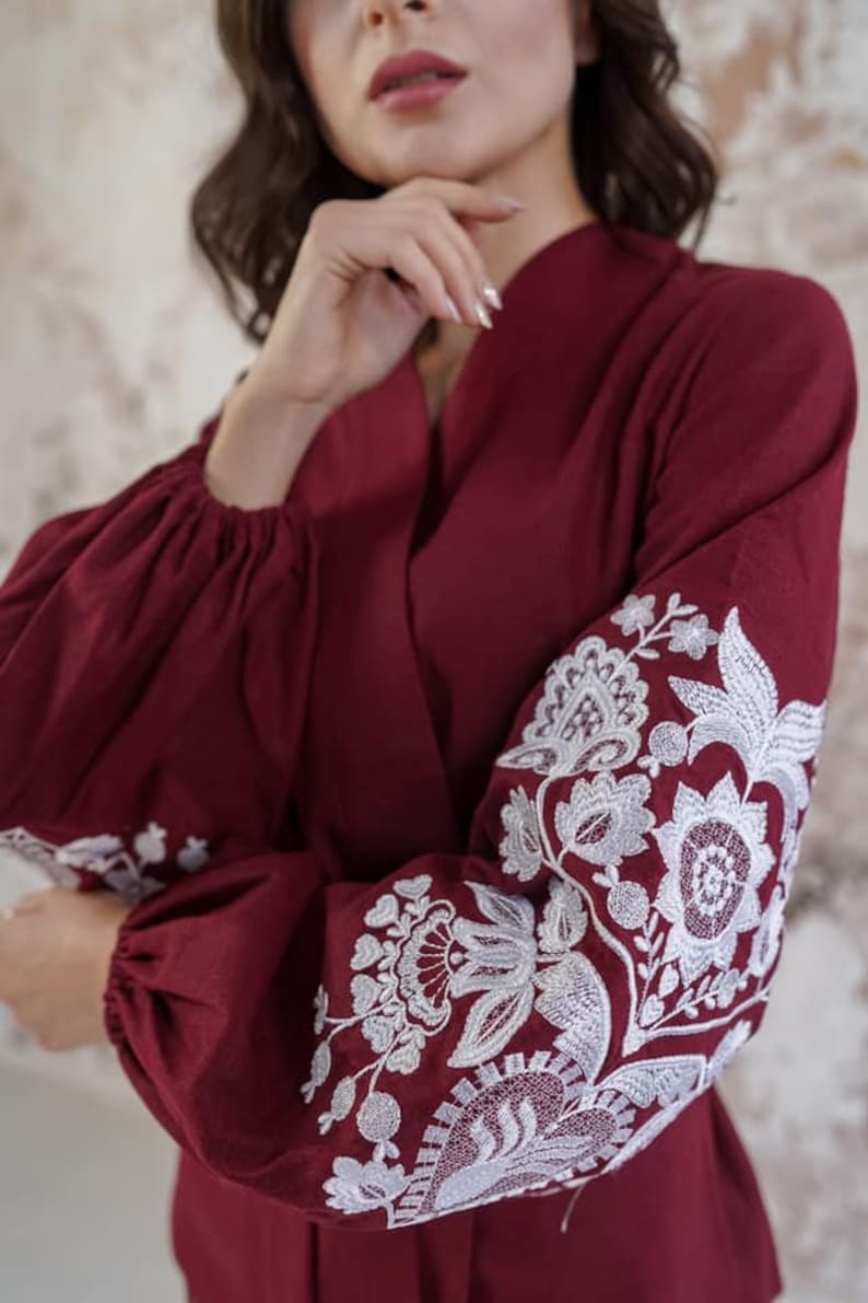 K&ouml;nnte beinhalten: Eine lang&auml;rmelige Bluse in Rotbraun mit wei&szlig;en, gestickten Blumenmustern an den &Auml;rmeln.