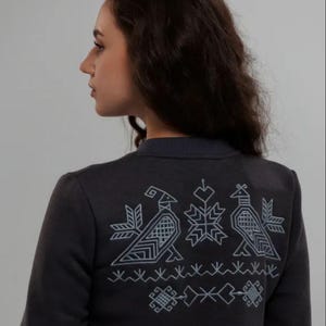 Puede incluir: Una sudadera gris oscuro con un dise&ntilde;o bordado blanco en la espalda. El dise&ntilde;o presenta dos p&aacute;jaros estilizados y un copo de nieve.