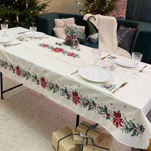 Puede incluir: Un mantel blanco con un diseño de acebo y poinsettia rojo y verde. El mantel está puesto para una cena navideña con platos, vasos y cubiertos.