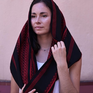 May include: A black head covering featuring intricate red geometric embroidery patterns along the borders and center panels. The fabric is draped over the head and shoulders, showcasing a detailed needlework design with repeating diamond motifs.