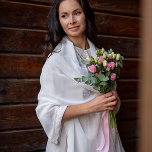Puede incluir: Una mujer con un chal y un vestido blancos, sosteniendo un ramo de flores rosas y verdes atado con una cinta rosa. El fondo es una pared de madera marr&oacute;n oscuro.
