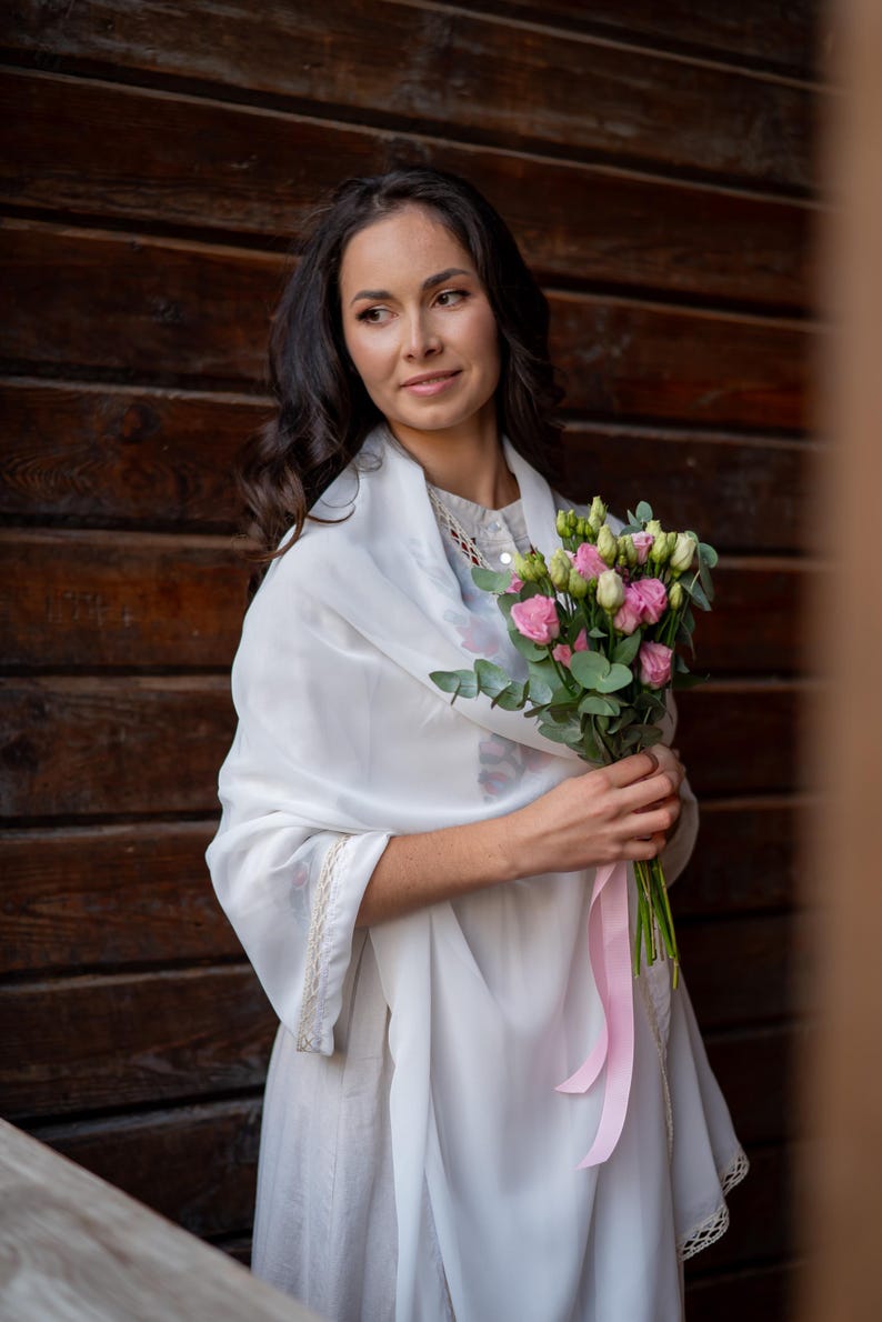 Puede incluir: Una mujer con un chal y un vestido blancos, sosteniendo un ramo de flores rosas y verdes atado con una cinta rosa. El fondo es una pared de madera marr&oacute;n oscuro.