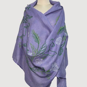 May include: A lilac shawl featuring green and white embroidered floral patterns. The shawl is draped over a mannequin, highlighting the detailed leaf and flower design. The fabric looks soft and lightweight, ideal for layering.