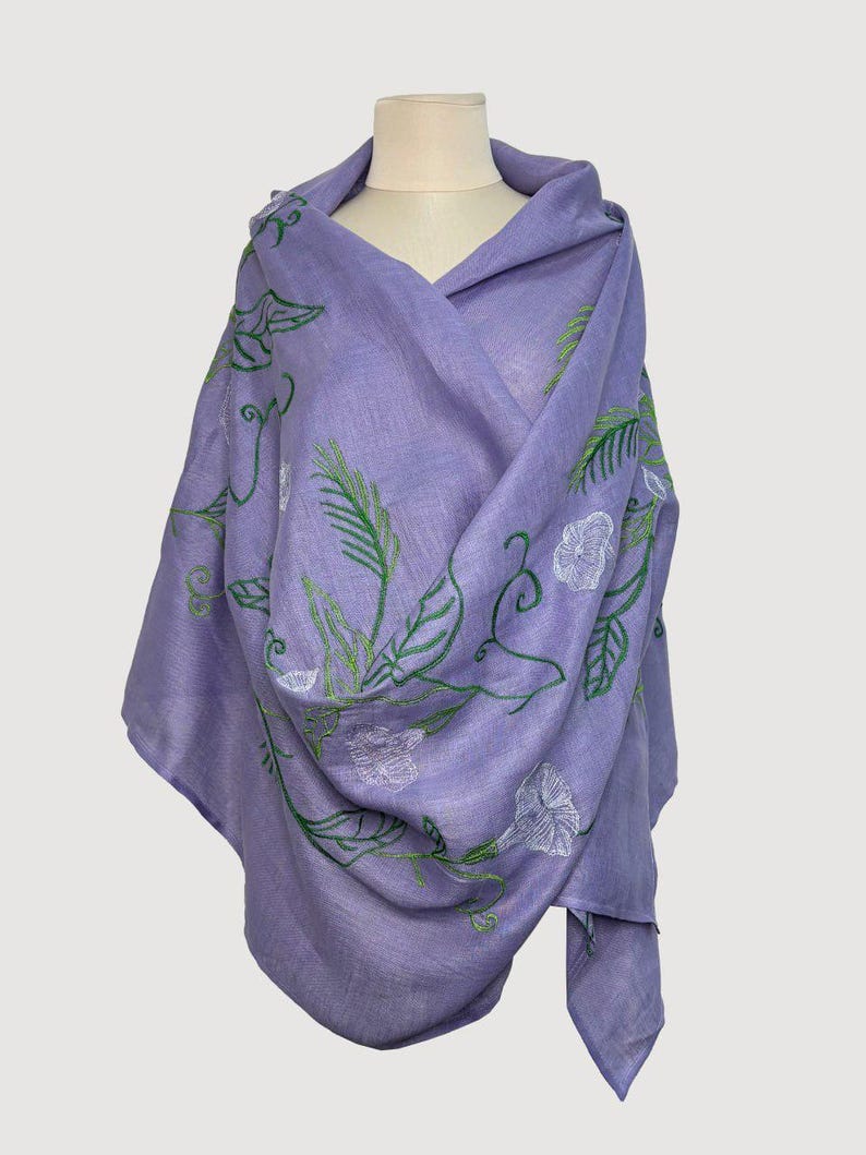 May include: A lilac shawl featuring green and white embroidered floral patterns. The shawl is draped over a mannequin, highlighting the detailed leaf and flower design. The fabric looks soft and lightweight, ideal for layering.