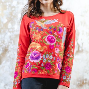May include: A red long-sleeved top adorned with colourful floral and bird embroidery. The design showcases a variety of flowers and birds in shades of pink, purple, blue, and orange. The top is a casual style.
