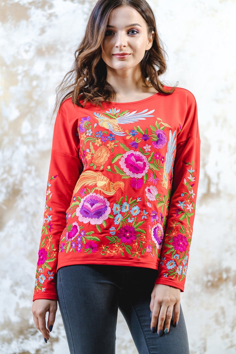 May include: A red long-sleeved top adorned with colourful floral and bird embroidery. The design showcases a variety of flowers and birds in shades of pink, purple, blue, and orange. The top is a casual style.