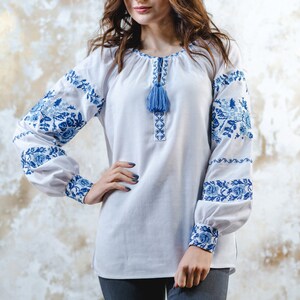 May include: A white embroidered blouse with blue floral patterns and a blue tassel. The blouse has long sleeves and a round neckline.