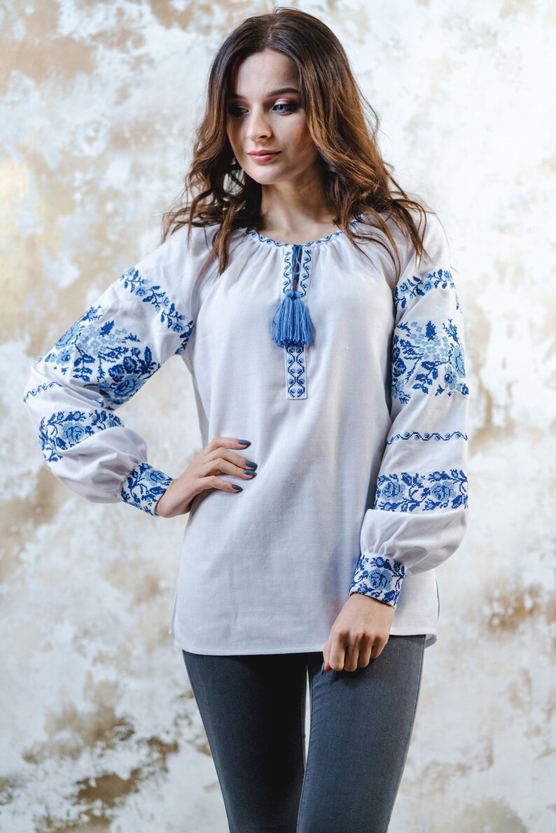 May include: A white embroidered blouse with blue floral patterns and a blue tassel. The blouse has long sleeves and a round neckline.