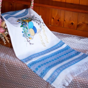 May include: White embroidered kitchen towel with blue trim and a design of a basket of Easter eggs. The text "Happy Easter" is embroidered in gold thread.