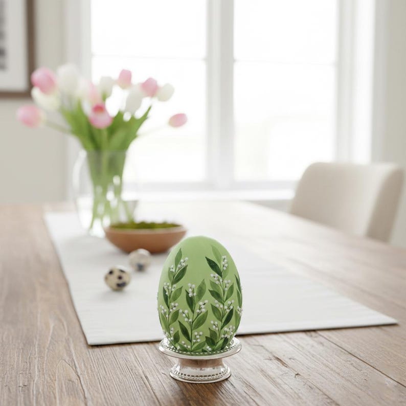 K&ouml;nnte beinhalten: Ein gr&uuml;nes Osterei mit floralem Design, pr&auml;sentiert in einem silbernen Eierbecher. Das Ei ist mit bemalten wei&szlig;en Blumen und gr&uuml;nen Bl&auml;ttern verziert. Eine Vase mit rosa und wei&szlig;en Tulpen und andere dekorative Eier befinden sich im Hintergrund.
