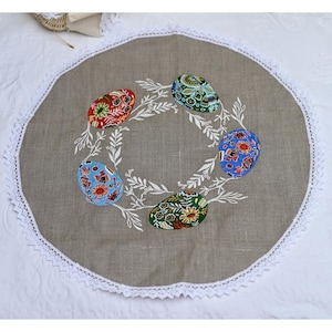 May include: A round, beige linen tablecloth with a white lace trim. The tablecloth features an embroidered design of five colourful Easter eggs surrounded by white leaves.
