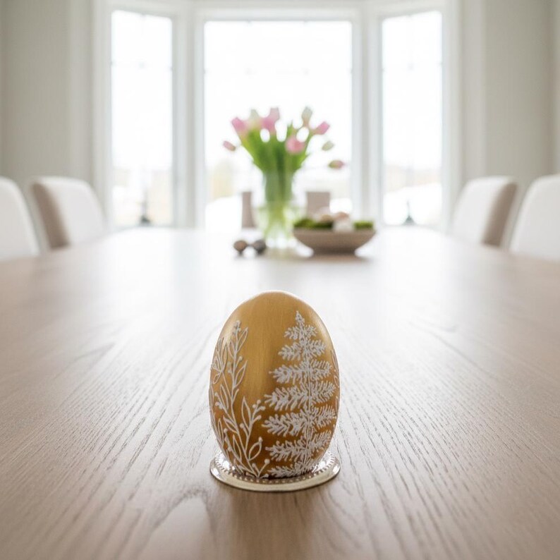 K&ouml;nnte beinhalten: Ein goldenes Ei mit wei&szlig; bemalten Blumen- und immergr&uuml;nen Designs, das auf einem silbernen Untersetzer ruht. Das Ei ist der Mittelpunkt auf einem hellbraunen Holztisch, mit einem verschwommenen Hintergrund eines Esszimmers.
