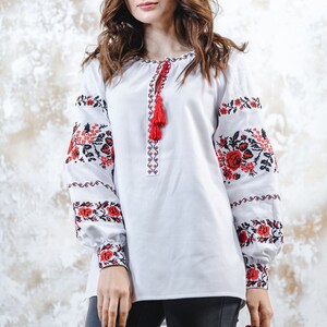 May include: A white embroidered blouse with red and black floral patterns on the sleeves. The blouse has a tassel detail at the neckline.