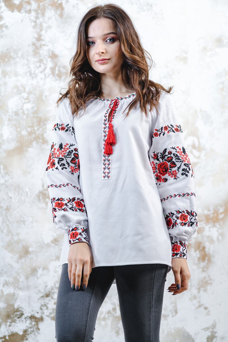 May include: A white embroidered blouse with red and black floral patterns on the sleeves. The blouse has a tassel detail at the neckline.