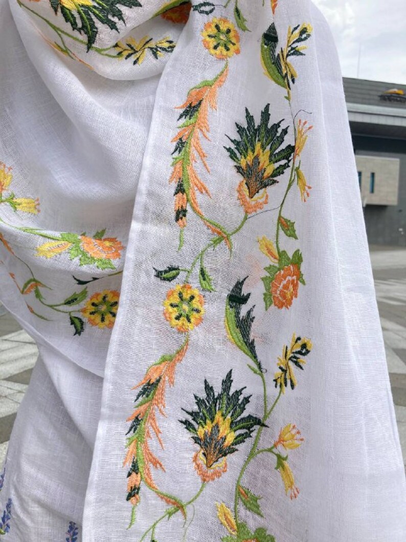 May include: White sheer scarf with embroidered floral design in shades of green, yellow, and orange.