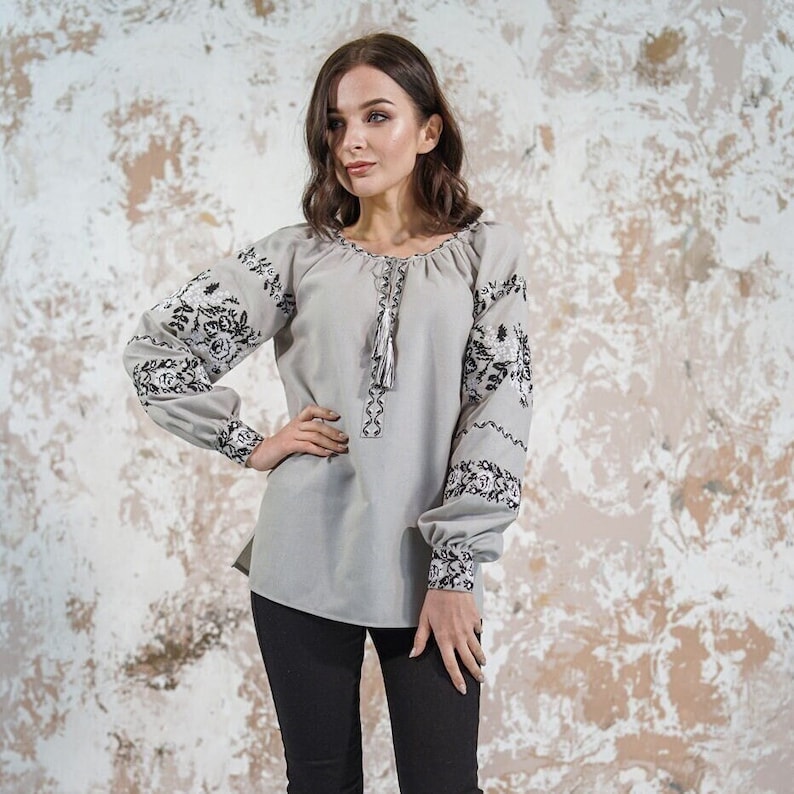 May include: A gray embroidered blouse with black floral designs on the sleeves. The blouse has a round neckline and a tassel detail at the front.