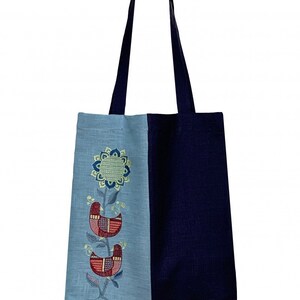 Puede incluir: Un bolso tote de dos tonos con asa y cuerpo azul marino, y un panel azul claro bordado con un dise&ntilde;o floral y de p&aacute;jaros. El bordado presenta hilos rojos, azules y verdes, creando un patr&oacute;n decorativo y colorido.