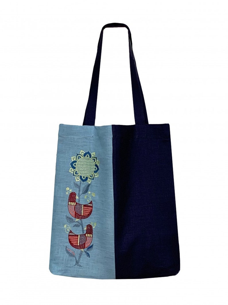 Puede incluir: Un bolso tote de dos tonos con asa y cuerpo azul marino, y un panel azul claro bordado con un dise&ntilde;o floral y de p&aacute;jaros. El bordado presenta hilos rojos, azules y verdes, creando un patr&oacute;n decorativo y colorido.