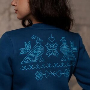Puede incluir: Una sudadera azul oscuro con un dise&ntilde;o bordado en azul claro en la espalda. El dise&ntilde;o presenta p&aacute;jaros estilizados y patrones geom&eacute;tricos.