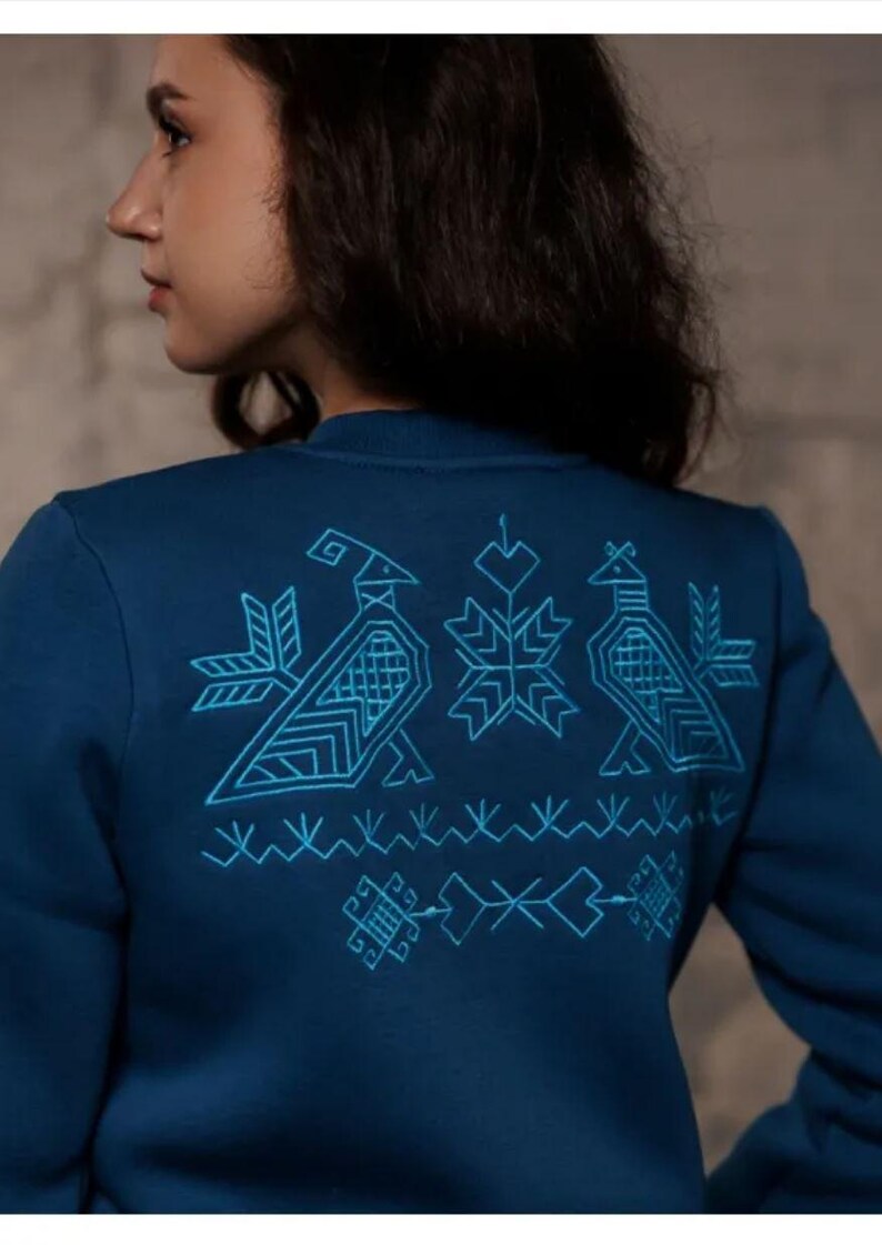 Puede incluir: Una sudadera azul oscuro con un dise&ntilde;o bordado en azul claro en la espalda. El dise&ntilde;o presenta p&aacute;jaros estilizados y patrones geom&eacute;tricos.