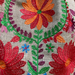 May include: Close-up of a beige fabric with colorful embroidered flowers. The flowers are red, orange, green, blue, and purple. The fabric has a woven texture.