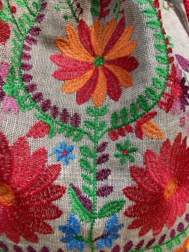 May include: Close-up of a beige fabric with colorful embroidered flowers. The flowers are red, orange, green, blue, and purple. The fabric has a woven texture.